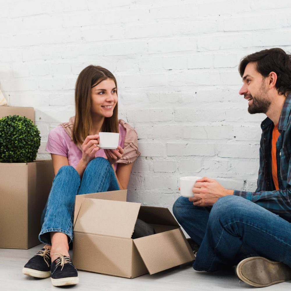 smiling-young-couple-sitting-floor-with-moving-cardboard-boxes-drinking-coffee(1)